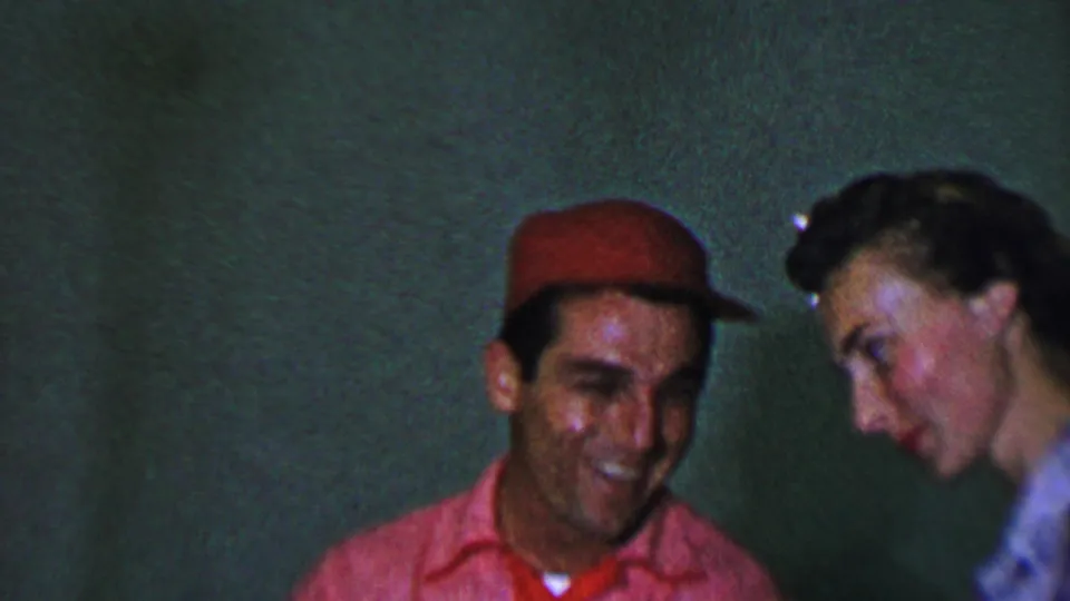 In this 1956 Dallas home-movie frame, the man in a red cap smiles while the woman leans close at his side in a dim indoor setting.