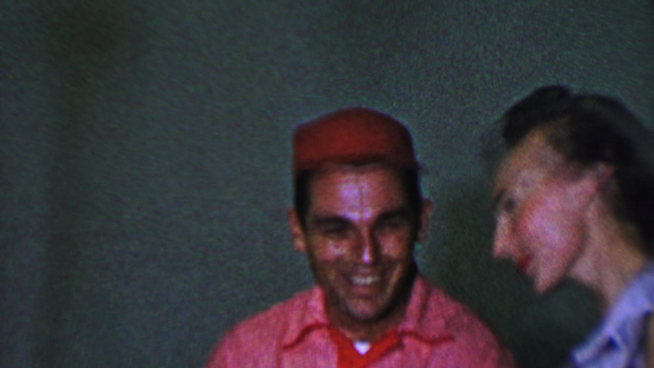 In this 1956 Dallas 8mm home-movie frame, a man in a red cap and pink shirt smiles while a woman beside him moves or laughs in soft blur against a plain green wall.