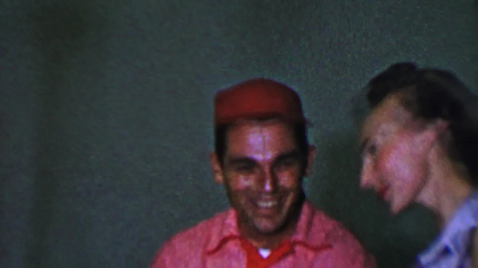 In this 1956 Dallas 8mm home-movie frame, a man in a red cap and pink shirt smiles while a woman beside him moves or laughs in soft blur against a plain green wall.