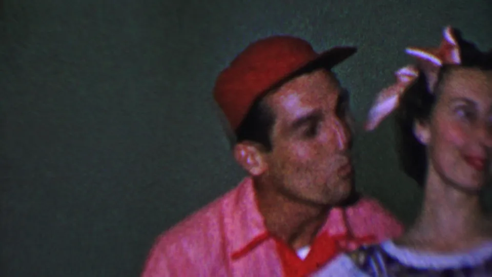 In this 1956 Dallas home-movie frame, a man in a red cap leans close to a woman wearing a light dress and hair bow, suggesting a playful affectionate moment.