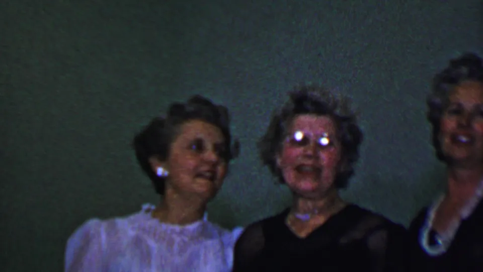 In this 1956 Dallas 8mm frame, women stand shoulder to shoulder near a green wall, with eyeglasses reflecting light and faces softened by motion and low exposure.
