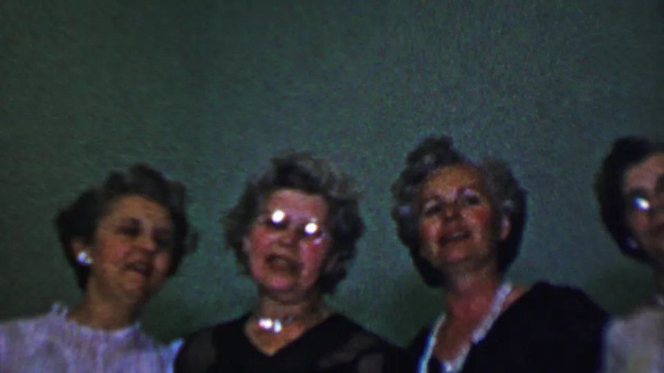 In this 1956 Dallas home-movie frame, four women appear close together against a green wall, smiling or singing toward the camera with one pair of eyeglasses reflecting the film light.