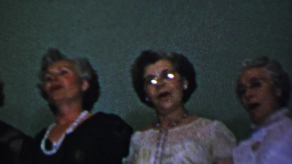 Dallas, Texas 1956 home-movie frame showing adult women singing in partial close-up, with jewelry, eyeglasses, and 1950s-style hair visible against a plain green wall.