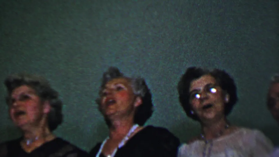 Dallas, Texas 1956 frame showing adult women in a small chorus, some wearing eyeglasses and jewelry, filmed in soft 8mm color against a plain indoor wall.