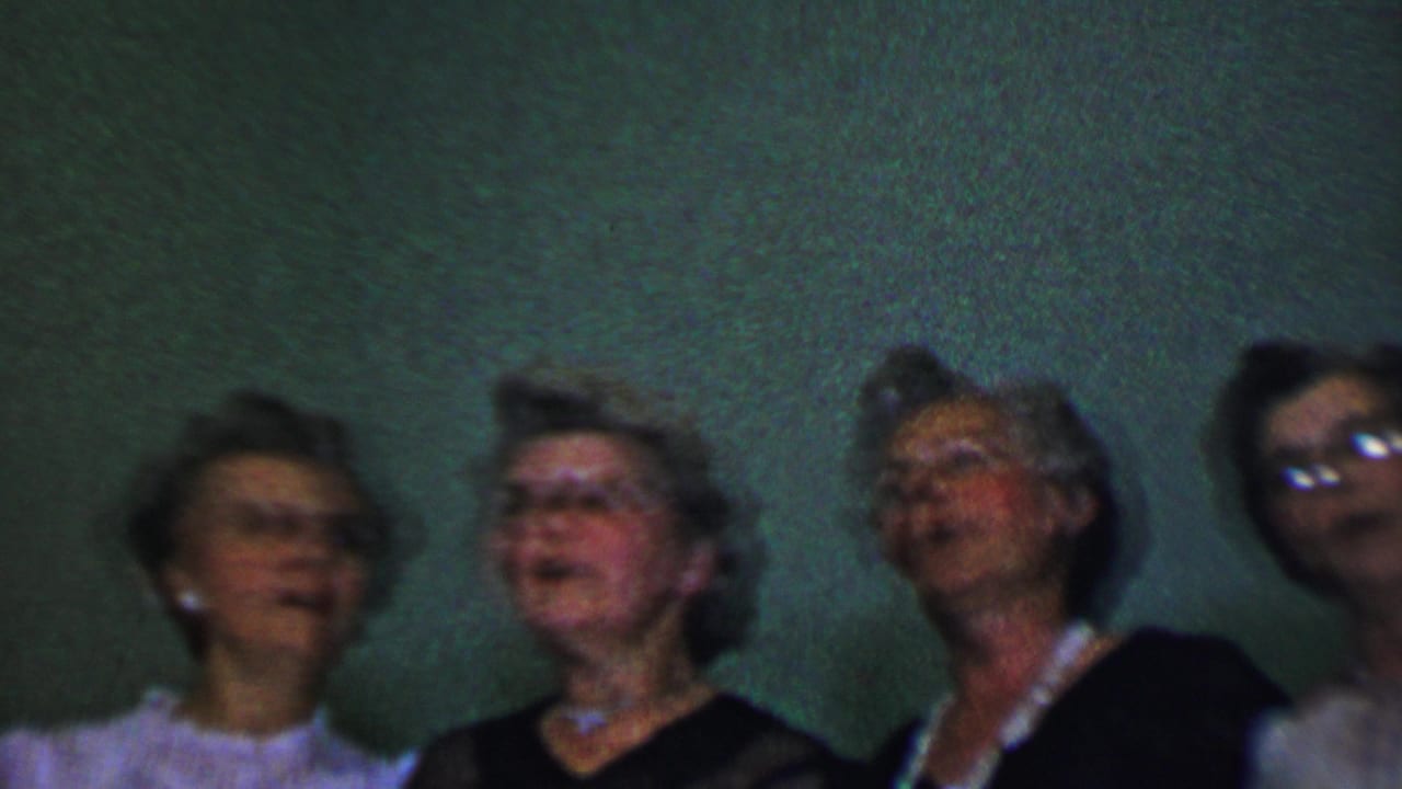 Adult women in dark and light formal dresses stand shoulder to shoulder against a textured green wall in Dallas home-movie footage from the supplied 1956 metadata.