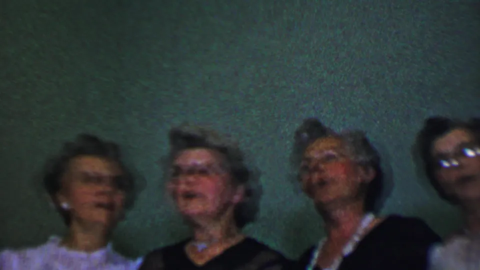 Adult women in dark and light formal dresses stand shoulder to shoulder against a textured green wall in Dallas home-movie footage from the supplied 1956 metadata.