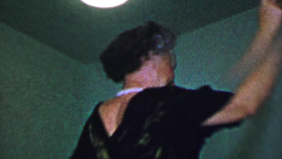 A woman in a dark dress with a light collar raises her arms while dancing in a dim Dallas interior recorded on 8mm film in 1956.