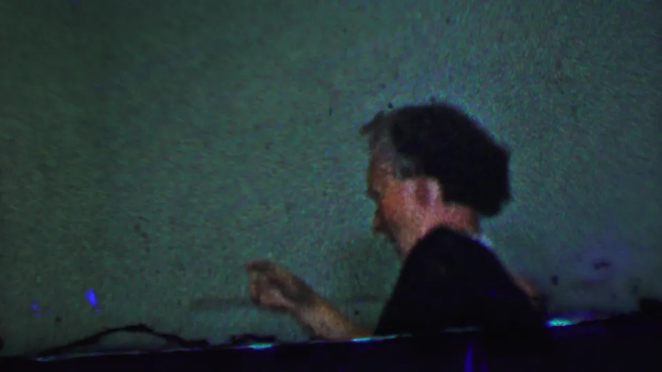 The woman appears in profile beside a green wall, gesturing as she moves through a dimly lit 1956 Dallas home-movie interior.