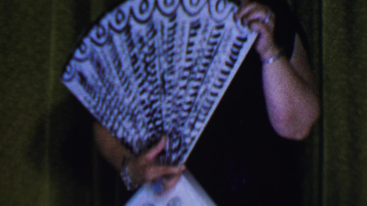 In a Dallas, Texas, 1956 home-movie frame, a woman on stage holds a patterned fan close to her face against a green curtain backdrop.
