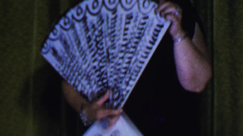 In a Dallas, Texas, 1956 home-movie frame, a woman on stage holds a patterned fan close to her face against a green curtain backdrop.