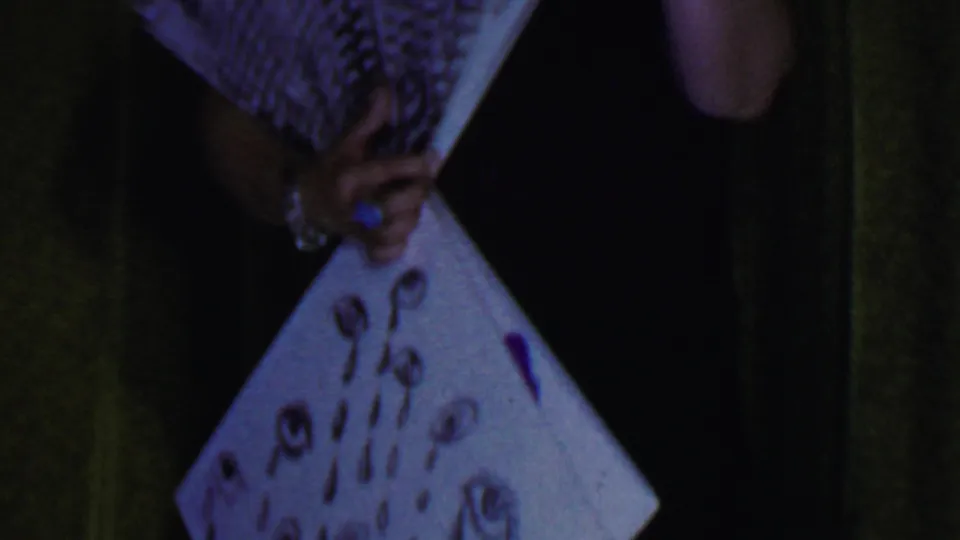 The stage performer holds a white fan or board with painted eyes and tear shapes while her face is partly obscured in the dim 8mm frame.