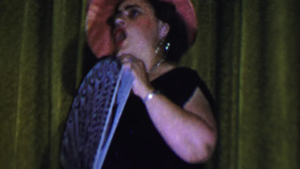 A woman in a pink hat and black dress appears to sing or emote while holding a patterned fan in front of a green curtain in the 1956 Dallas footage.
