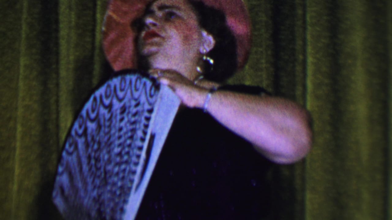 In supported 1956 Dallas footage, a woman in a pink hat and dark dress holds a large patterned fan while performing against a green curtain.