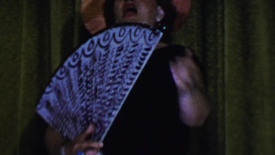 The performer, wearing a pink hat and dark dress, is partly obscured by a large open fan under stage lighting in the Dallas home-movie clip.