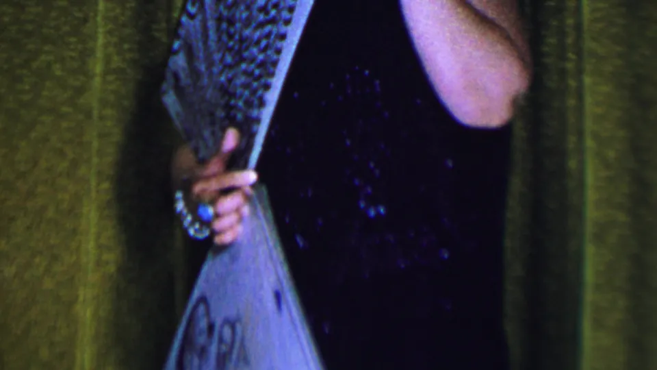A closer 8mm frame shows the woman on stage with the fan lowered across her dark dress, with jewelry visible on her hand and wrist.
