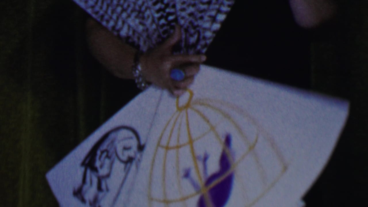 1956 Dallas 8mm frame showing a performer's hand with bracelet and blue ring holding a white fan decorated with a golden cage and colorful figures.
