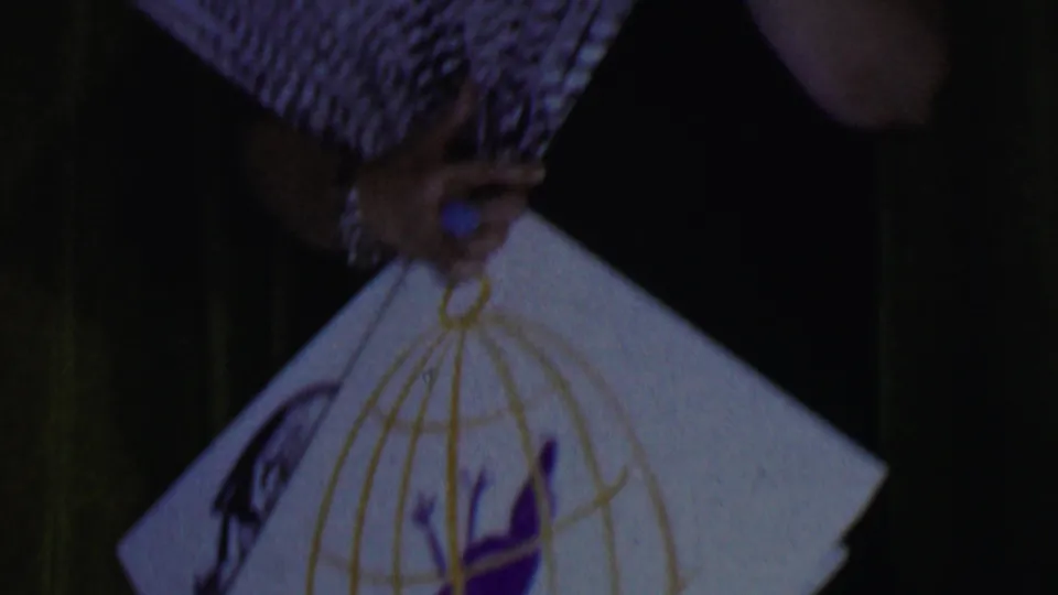 1956 Dallas 8mm frame with a close, soft-focus view of a decorative white fan held by a performer wearing jewelry against a dark stage background.