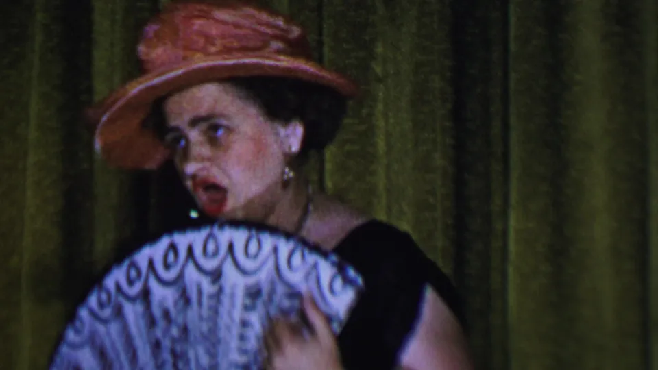 1956 Dallas 8mm frame showing a woman performer in a pink wide-brimmed hat, black dress, hoop earrings, and red lipstick singing behind an open white fan.