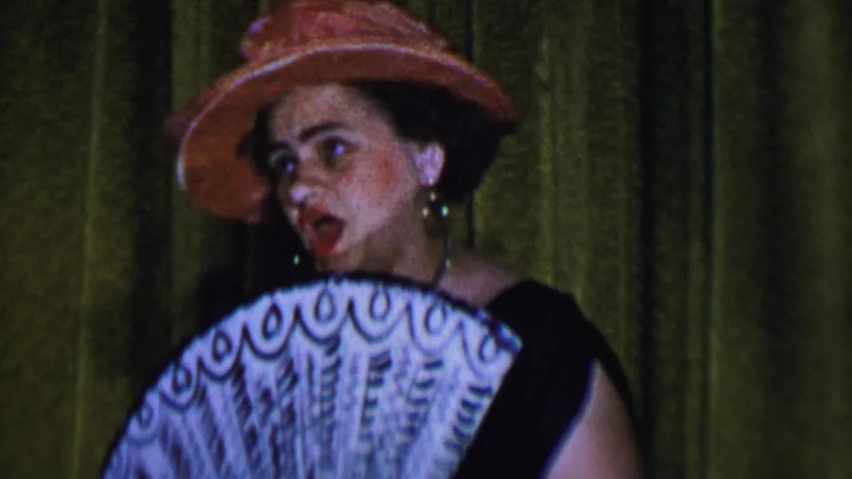 1956 Dallas 8mm frame showing the woman singer turned to one side, wearing a pink hat and hoop earrings while holding an open white fan on stage.