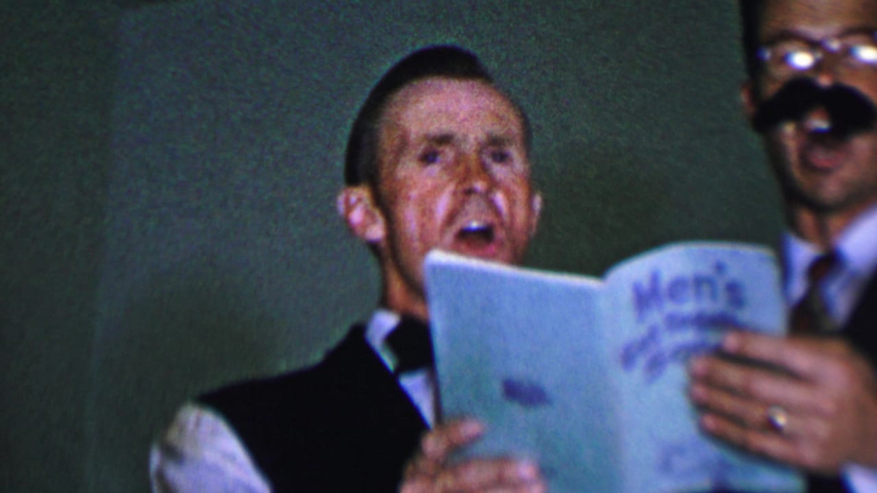 In the supplied 1956 Dallas home-movie context, men in vests and bow ties sing from a blue Men's Get-Together Songs booklet during an indoor gathering.