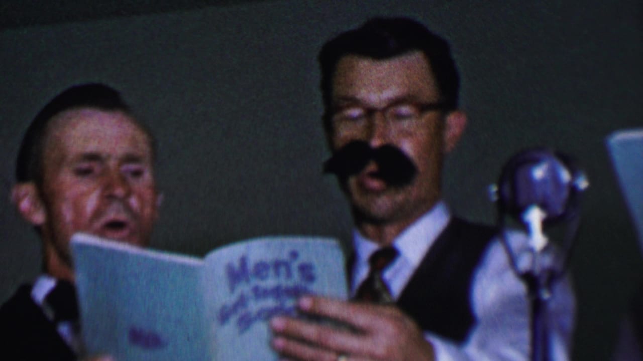 A man with a fake mustache sings beside another man holding a blue songbook near a microphone.