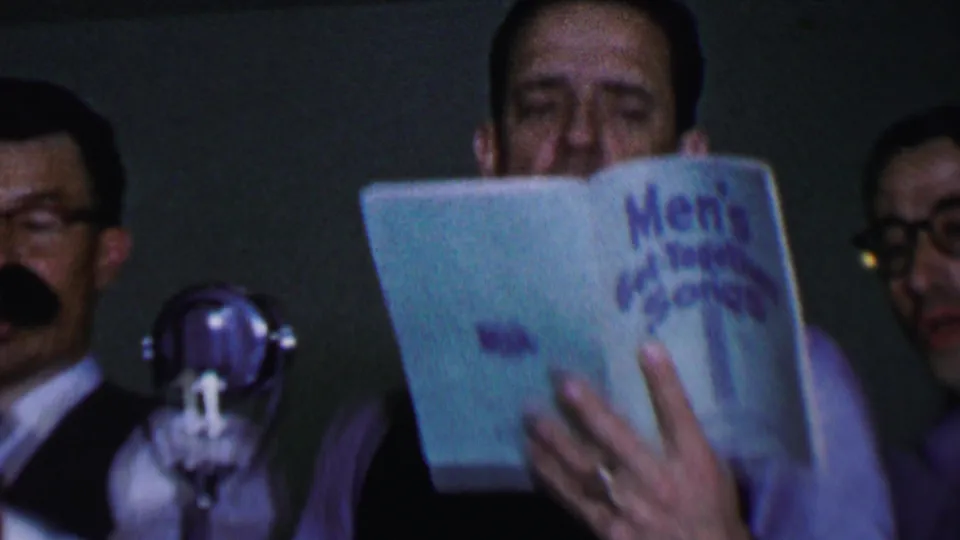 A close, low-light 8mm frame shows a man singing from the blue Men's Get-Together Songs booklet, with other men partly visible at the sides and a microphone in front.
