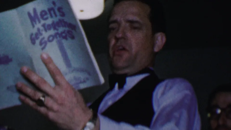 A man in formal vest and bow tie sings from the Men's Get-Together Songs booklet in a dim indoor frame, with another partially visible face at the lower right.