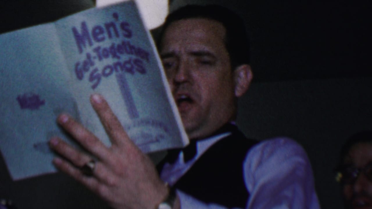 In 1956 Dallas home-movie footage, a man in a bow tie and vest sings from a Men's Get-Together Songs book in a dim indoor room.