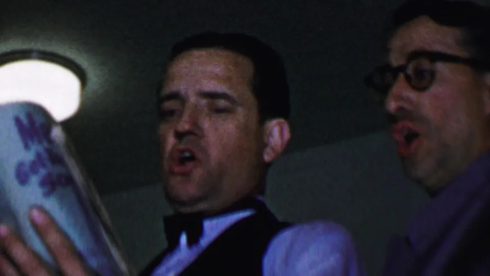 Two men appear to sing indoors in the Dallas 1956 footage, with one man in formalwear reading from a songbook and another man with glasses at the side.