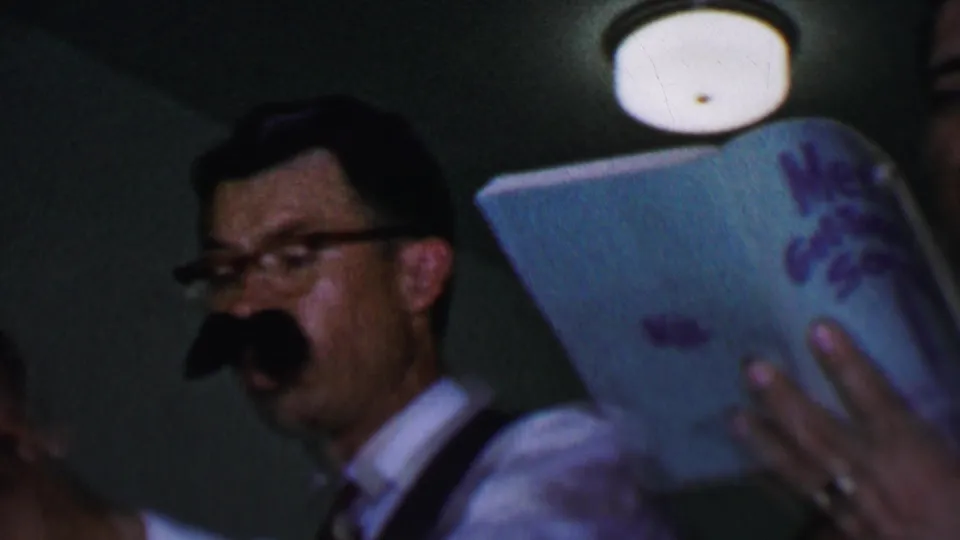 A man wearing glasses and a fake mustache stands near a Men's Get-Together Songs book and microphone during the 1956 Dallas indoor gathering.