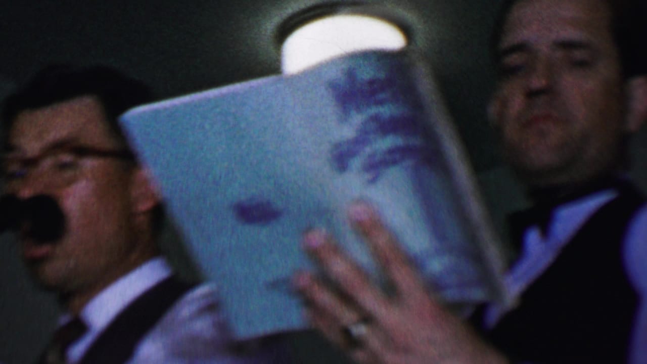 In supported 1956 Dallas home-movie footage, adult men in dress shirts, vests, and bow ties appear to sing while one holds an open booklet close to the camera.