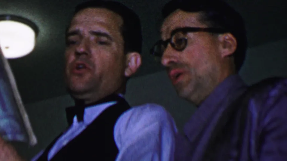 Three adult men in formal shirts, dark vests, and bow ties appear to sing from booklets in a dim private interior, with one man wearing glasses.