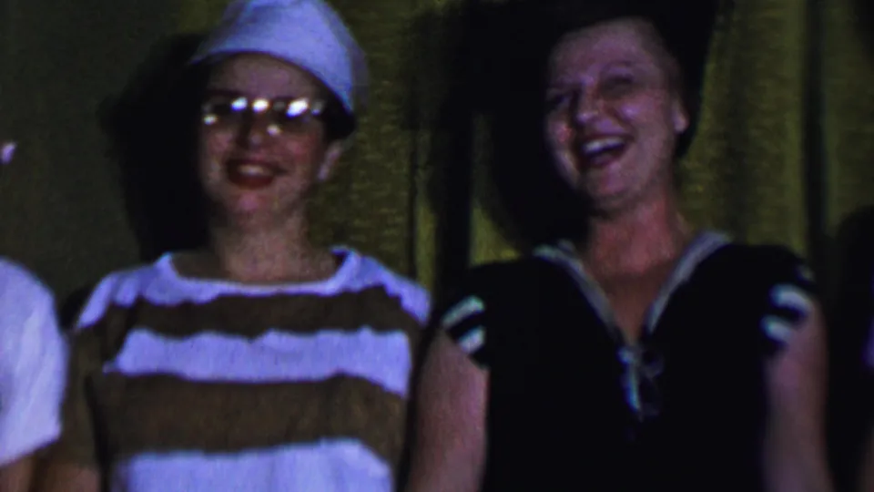 Adult women in mid-century clothing laugh and look toward the camera or nearby performers, with striped tops, light hats, glasses, and a dark interior background visible.