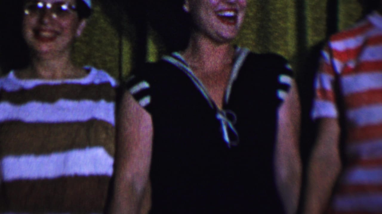 Dallas 1956 home-movie frame showing women standing close together indoors, with striped clothing, eyeglasses, and a dark sailor-style dress visible in dim light.