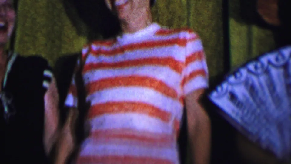Dallas 1956 home-movie frame showing a woman in a red-and-white striped shirt beside partially visible companions and a decorative fan.