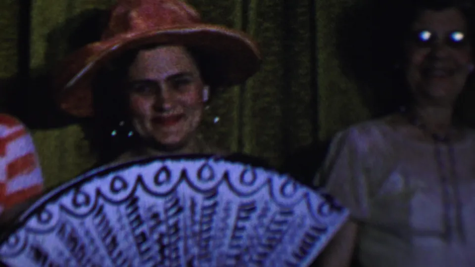 Dallas 1956 home-movie frame showing a woman in a wide-brimmed hat holding a decorative fan, with another smiling woman in glasses nearby.