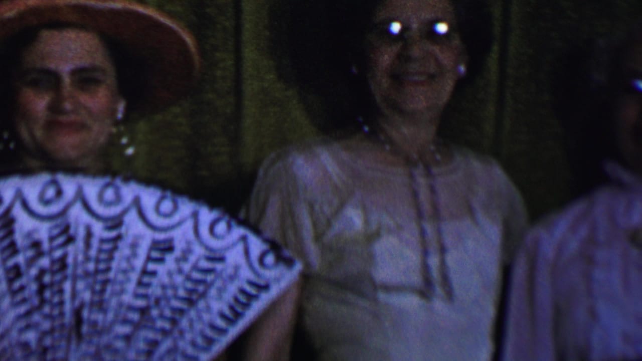 In this 1956 Dallas home-movie frame, two adult women face the camera, with one wearing a wide-brimmed hat and patterned fan-like fabric and another in glasses before dark green curtains.