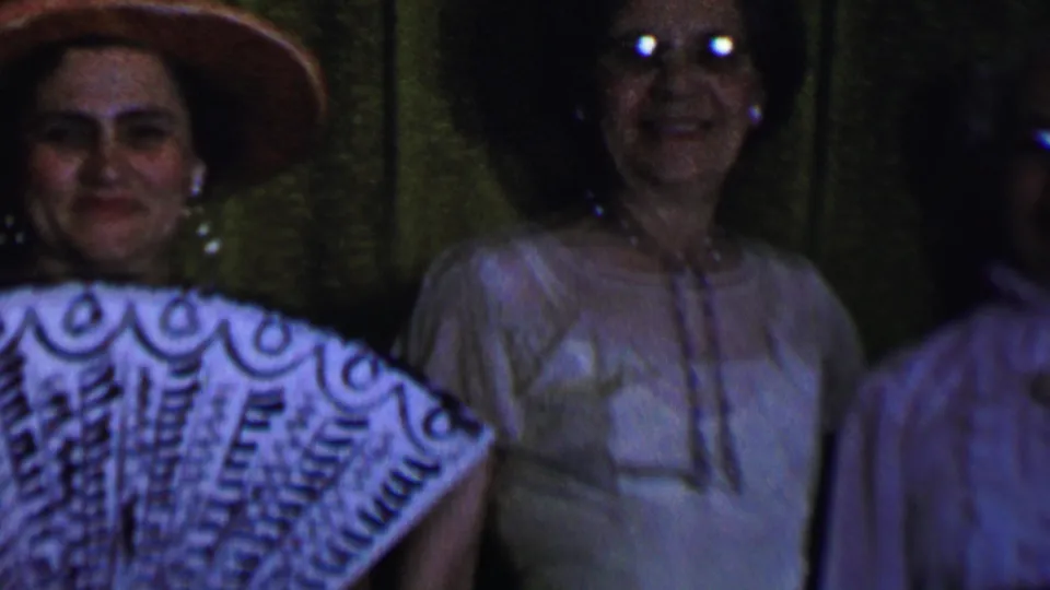 In this 1956 Dallas home-movie frame, two adult women face the camera, with one wearing a wide-brimmed hat and patterned fan-like fabric and another in glasses before dark green curtains.