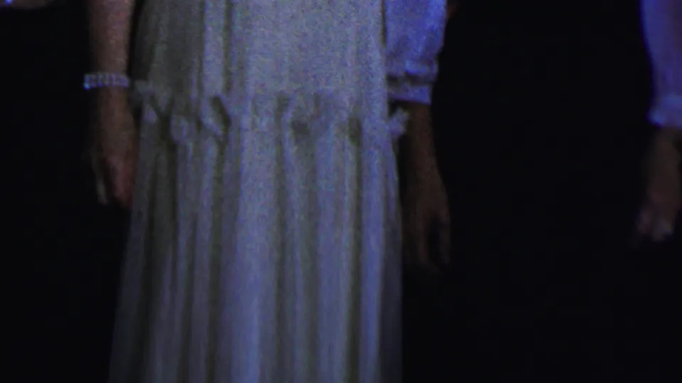 A cropped 1956 Dallas home-movie frame shows adult women from shoulders to knees, including a pale dress with gathered detail and a dark dress with a light sleeve visible.