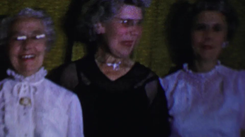In this 1956 Dallas home-movie frame, three adult women stand close together before green curtains, with visible glasses, jewelry, a dark dress, and a pale blouse or dress.