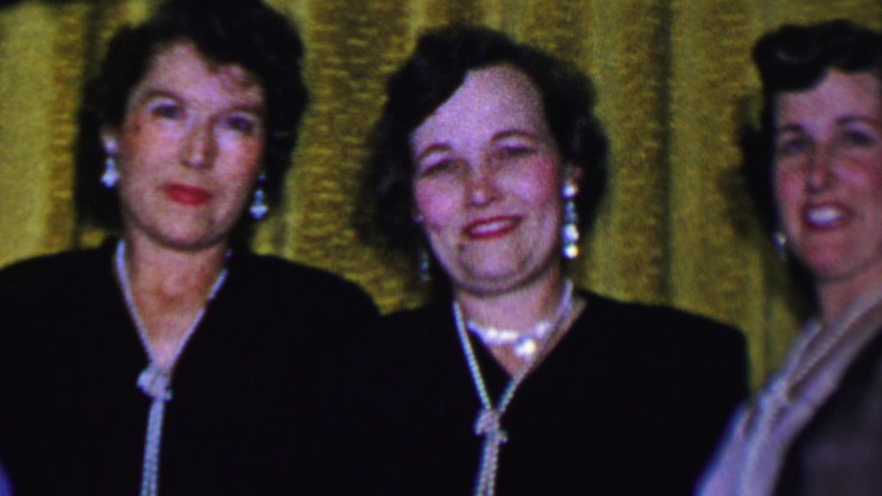Adult women in Dallas, Texas, in 1956 appear dressed for a formal social gathering, wearing dark dresses and jewelry against a gold curtain backdrop.