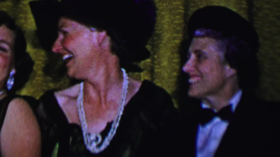 Adults at a Dallas social gathering in 1956 laugh and look sideways, with formally dressed women and a partially visible man in a tuxedo near the gold curtain.