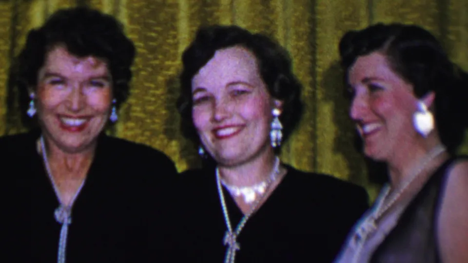 Three adult women at the 1956 Dallas gathering smile toward the camera, wearing formal dark dresses, jewelry, and styled hair before the gold curtain backdrop.