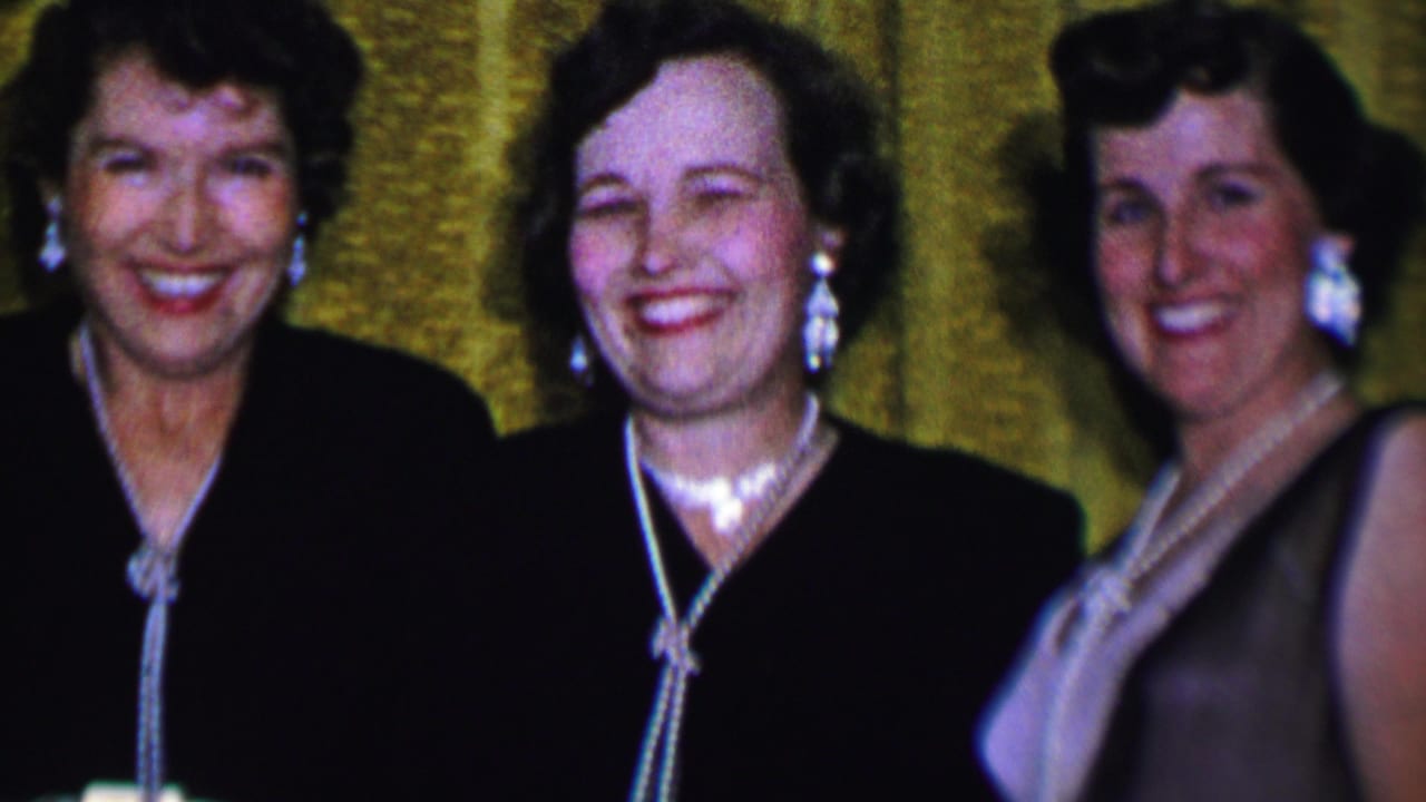 Three women in Dallas, Texas, 1956 home-movie footage stand close together in dark formal dresses with pearl jewelry, smiling before a textured gold curtain.