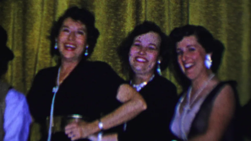Women in 1956 Dallas home-movie footage laugh and pose together in formal dresses, pearl necklaces, and earrings against a gold curtain backdrop.