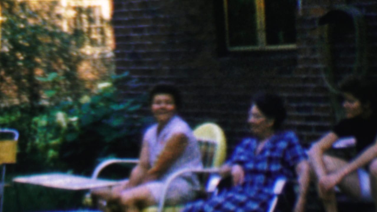 Adults relax on metal lawn chairs near a brick porch and green foliage.