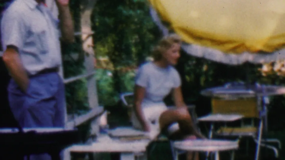 A man in a light shirt and blue trousers stands near a seated woman in a light-colored outfit, with patio chairs, a small table, greenery, and a yellow umbrella visible in the 1961 Covington home-movie scene.