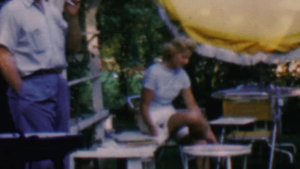 The frame shows a casually dressed man standing near a seated woman on a porch or patio, surrounded by metal furniture, a yellow umbrella, steps, and dense greenery.