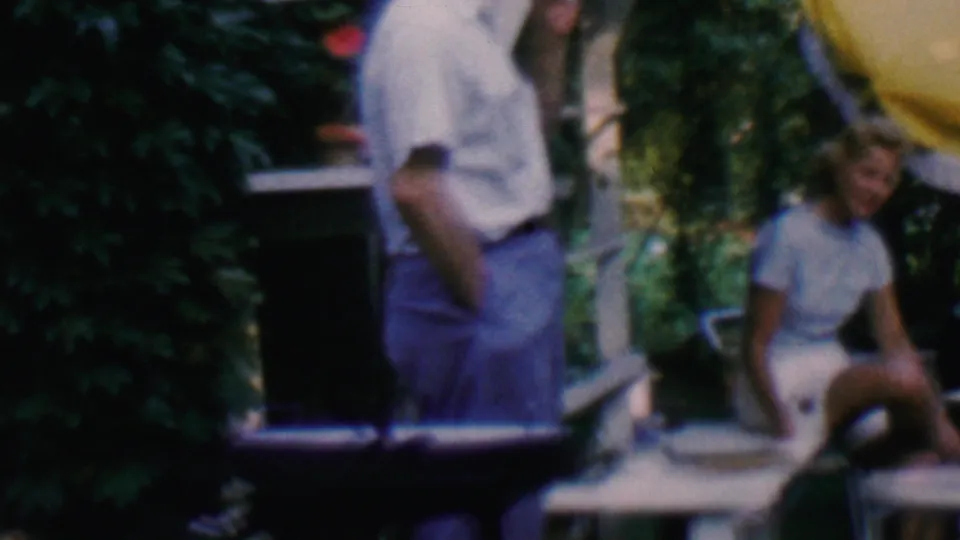Near the end of the clip, the 8mm frame holds on a standing man and seated woman by metal patio chairs, greenery, porch steps, and the edge of a yellow umbrella.
