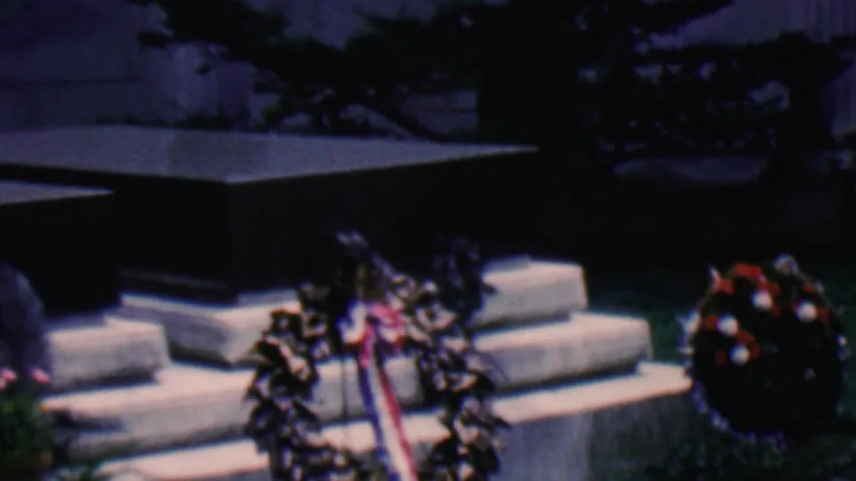 Covington, Kentucky, 1961 frame with a ribboned wreath and a round floral arrangement beside a dark stone monument and low steps.